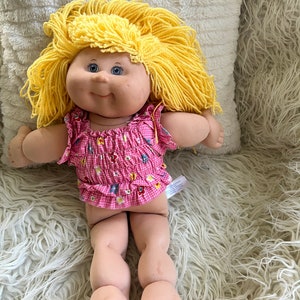 May include: A Cabbage Patch Kid doll with yellow yarn hair, wearing a pink and white gingham dress with floral accents. The doll is sitting on a white fluffy surface.