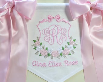 May include: A white banner with pink embroidery of a floral wreath, a monogram, and the name "Gina Elise Ross". The banner is decorated with two pink satin bows.
