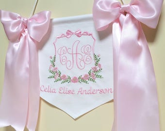 May include: A white fabric banner with pink embroidered floral accents and the name "Celia Elise Anderson". Two pink satin bows are attached to the banner.