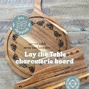 May include: Two wooden charcuterie boards with handles. The round board has a decorative leaf design and the text "Lay the table with the fancy shit." The rectangular board has the same text. Both boards are made of light brown wood.