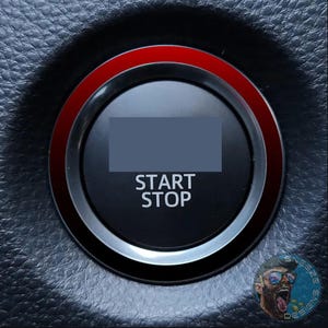 May include: A circular black engine start stop button featuring a prominent red ring around the outer edge. The button has the words START STOP printed in white capital letters in the center, set against a textured dark gray background.