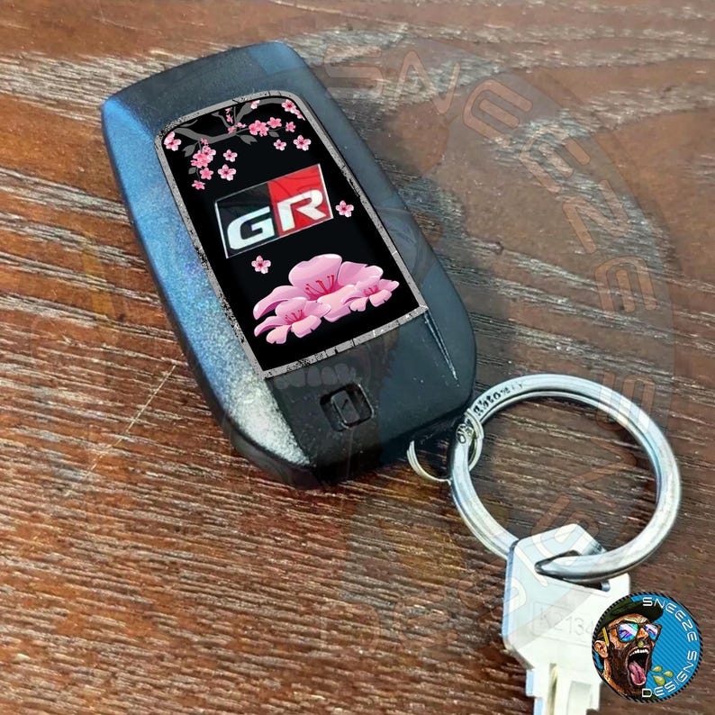 Puede incluir: Un llavero de coche negro con un dise&ntilde;o de flor de cerezo y las letras "GR" en rojo. El llavero est&aacute; unido a un llavero plateado.