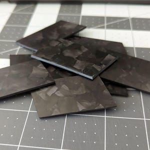 May include: A collection of rectangular, dark gray carbon fiber pieces with a textured pattern. The pieces are scattered on a gray and white grid surface, suggesting a crafting or industrial theme.