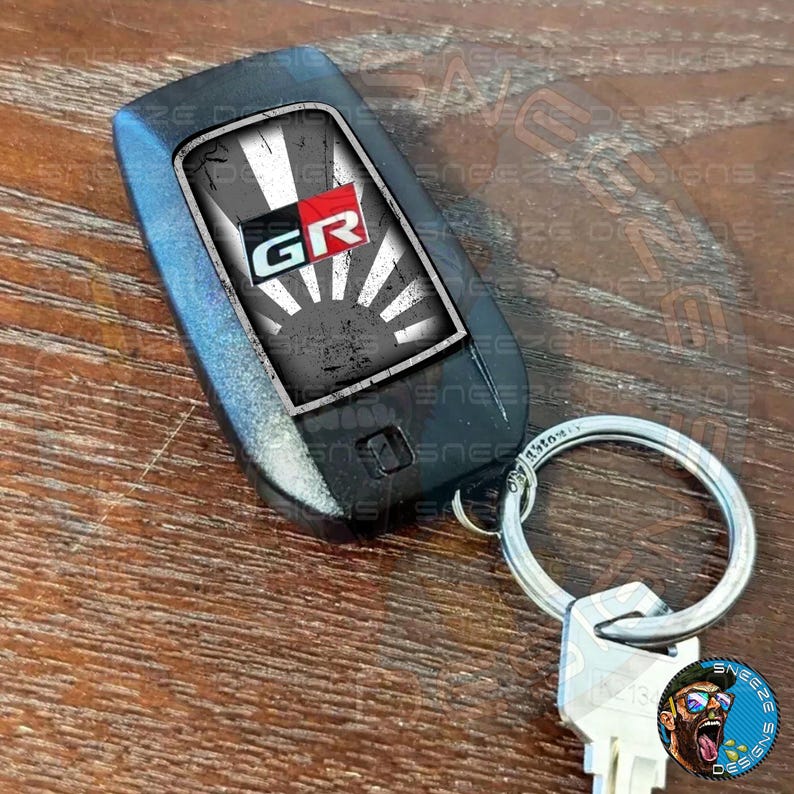 Puede incluir: Un llavero de coche negro con un dise&ntilde;o gr&aacute;fico blanco y negro que presenta las letras "GR" en rojo y blanco. El llavero est&aacute; unido a un llavero plateado.