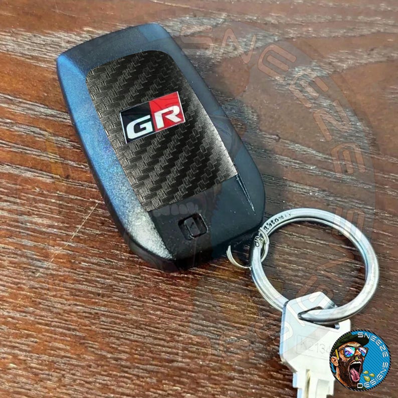 Puede incluir: Un llavero de coche negro con un patr&oacute;n de fibra de carbono y un logotipo "GR" rojo y blanco. El llavero est&aacute; unido a un llavero plateado.