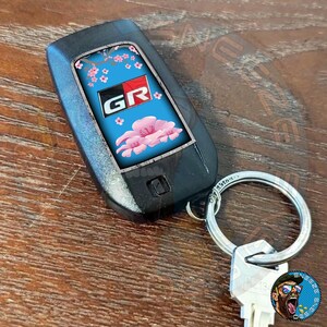 Puede incluir: Un llavero de coche negro con un dise&ntilde;o azul y rosa con un patr&oacute;n de flor de cerezo y las letras "GR" en rojo y blanco. El llavero est&aacute; unido a un llavero plateado.