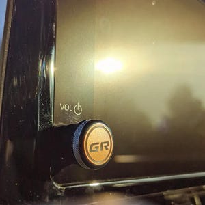 May include: Close-up of a car's infotainment system. The volume knob is black with a gold-colored center featuring the letters "GR". The screen displays a bright, blurred background. The word "VOL" and a power symbol are visible.