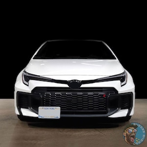 May include: A white car viewed from the front, featuring a black grille with a small red accent, sharp angular headlights, and a New York license plate mounted on the lower bumper. The vehicle has a sleek hood design and a black emblem in the center.
