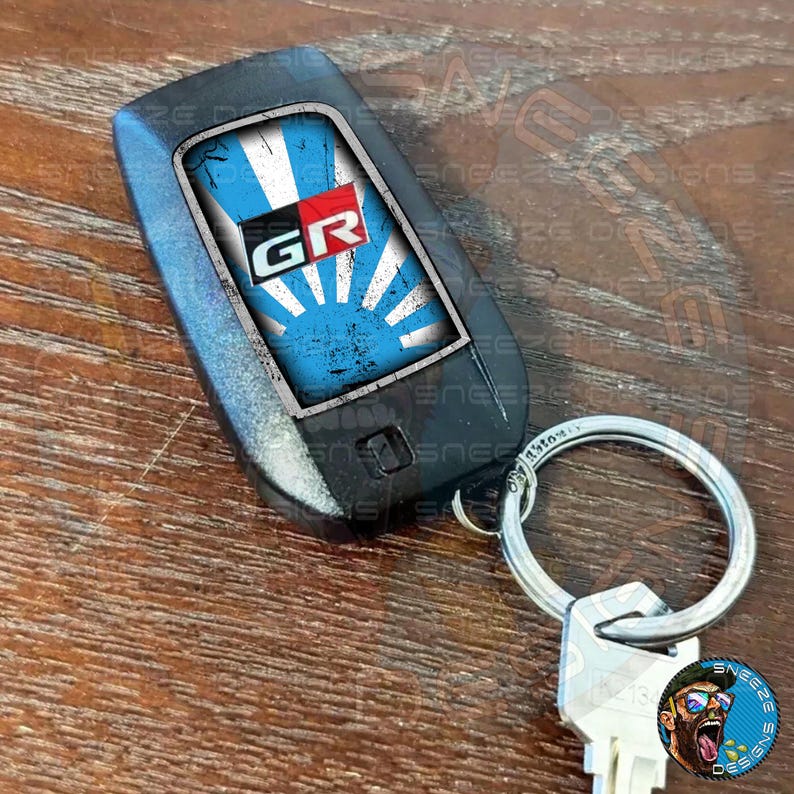 Puede incluir: Un llavero de coche negro con un dise&ntilde;o gr&aacute;fico azul, blanco y rojo que presenta las letras "GR" y un sol estilizado. El llavero est&aacute; unido a un llavero plateado.
