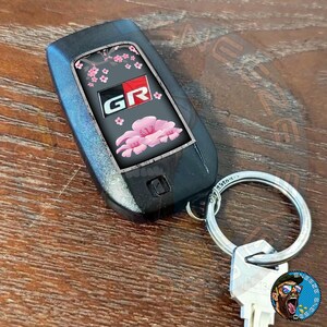 Puede incluir: Llavero de coche negro con un dise&ntilde;o de flor de cerezo rosa y blanco y las letras "GR" en rojo. El llavero est&aacute; unido a un llavero plateado.