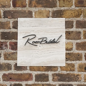 Op de afbeelding: Een vierkant, gebroken wit marmeren bord met de zwarte tekst "Roco Bridal" gemonteerd op een bakstenen muur. Het bord heeft een getextureerd oppervlak met subtiele golfpatronen.