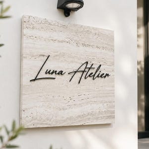 Puede incluir: Un letrero cuadrado de piedra travertino beige con el texto negro Luna Atelier en fuente cursiva. El letrero está montado en una pared blanca debajo de una lámpara de pared negra. La piedra tiene una textura natural con vetas horizontales.