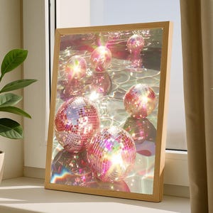 May include: A framed print featuring multiple disco balls reflecting light. The balls are pink and silver, set against a watery background. The wooden frame adds a natural touch to the vibrant, colorful scene.