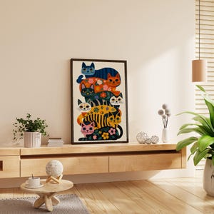 May include: A framed art print featuring a stack of stylized cats in various colors and patterns. The cats include blue, orange, green, yellow, and pink, with floral and striped designs. The artwork is displayed on a light-colored wall.