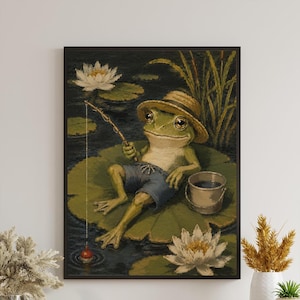 May include: A framed wall art print depicting a green frog wearing a straw hat and blue shorts, sitting on a large lily pad in a dark pond. The frog holds a fishing rod with a red and white bobber in the water, next to a small bucket and white lilies.