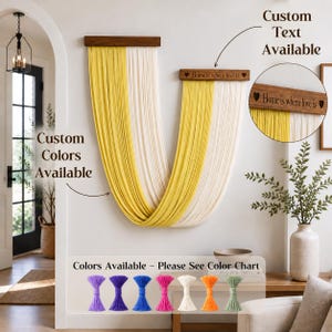 May include: A wall hanging featuring two wooden bars with long, draped yellow and cream-colored fabric strands. The smaller wooden bar is engraved with the text "Home is where love is" flanked by two heart symbols. A color chart shows six tassels.