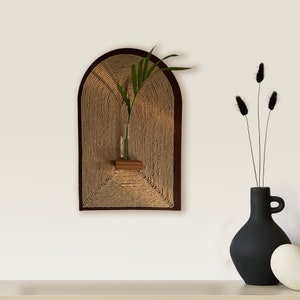 Pode incluir: Uma tapeçaria de parede de corda marrom com uma pequena prateleira de madeira e um vaso de vidro com uma planta verde. A tapeçaria de parede tem a forma de um arco.