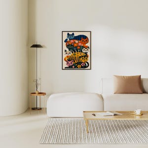 May include: A framed art print featuring a stack of stylized cats in various colors, including blue, orange, and yellow. The cats are decorated with floral patterns and sit against a cream background. The artwork is displayed on a white wall.