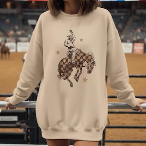 May include: Sand-colored sweatshirt featuring a rodeo-themed graphic. The design depicts a cowboy riding a bucking bronco with a checkered pattern, accompanied by star accents. The sweatshirt has a crew neck and long sleeves, perfect for casual wear.