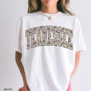Puede incluir: Camiseta blanca con la palabra "TENNESSEE" en un dise&ntilde;o de camuflaje en el pecho. El texto est&aacute; en letras de bloque con una ligera curva. La camiseta tiene mangas cortas y cuello redondo.