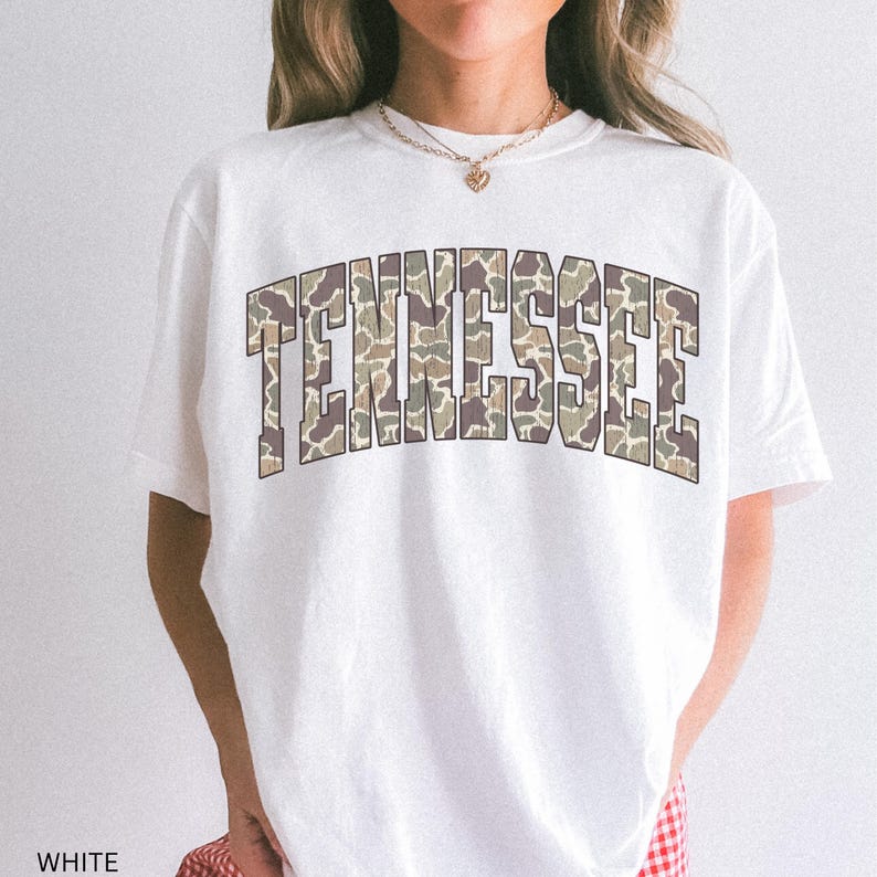 Puede incluir: Camiseta blanca con la palabra "TENNESSEE" en un dise&ntilde;o de camuflaje en el pecho. El texto est&aacute; en letras de bloque con una ligera curva. La camiseta tiene mangas cortas y cuello redondo.