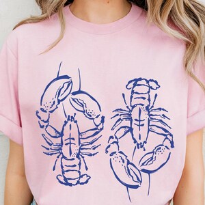以下が含まれることがあります： 明るいピンク色のTシャツ。ネイビーブルーのロブスターのイラストが2つ描かれています。Tシャツは半袖で、クルーネックです。「BLOSSOM」の文字がTシャツの下部にプリントされています。