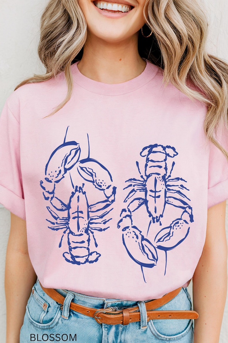 以下が含まれることがあります： 明るいピンク色のTシャツ。ネイビーブルーのロブスターのイラストが2つ描かれています。Tシャツは半袖で、クルーネックです。「BLOSSOM」の文字がTシャツの下部にプリントされています。