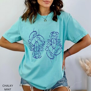 以下が含まれることがあります： 明るいブルーのTシャツ。濃いブルーのロブスターのグラフィックが描かれています。半袖でゆったりとしたフィット感です。画像左下には「CHALKY MINT」の文字がプリントされています。