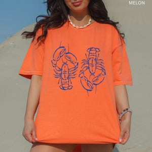 以下が含まれることがあります： 青いロブスターのグラフィックが入ったオレンジ色のTシャツ。Tシャツは半袖とクルーネックです。「MELON」の文字が右上隅に印刷されています。