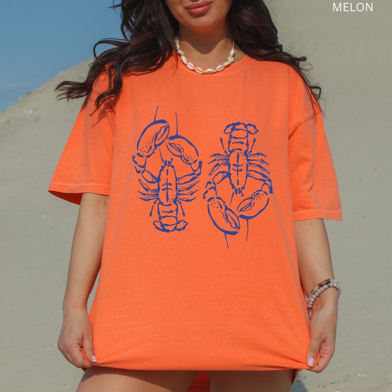 以下が含まれることがあります： 青いロブスターのグラフィックが入ったオレンジ色のTシャツ。Tシャツは半袖とクルーネックです。「MELON」の文字が右上隅に印刷されています。