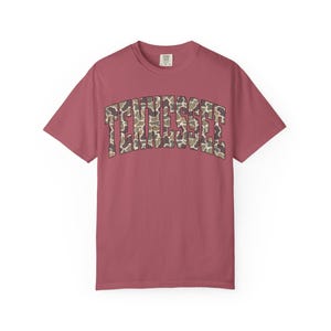 Puede incluir: Camiseta de manga corta color rosa polvoriento con la palabra "TENNESSEE" arqueada en el pecho. El texto presenta un estampado de camuflaje en marr&oacute;n, beige y verde. La camiseta tiene cuello redondo y corte holgado.