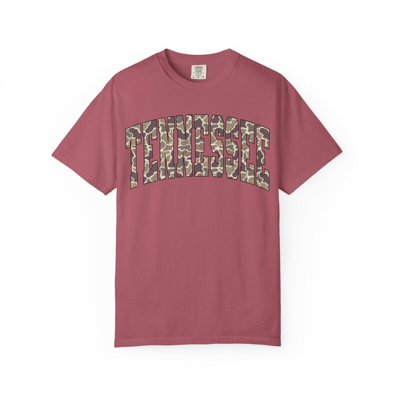 Puede incluir: Camiseta de manga corta color rosa polvoriento con la palabra "TENNESSEE" arqueada en el pecho. El texto presenta un estampado de camuflaje en marr&oacute;n, beige y verde. La camiseta tiene cuello redondo y corte holgado.