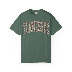 Puede incluir: Camiseta verde con la palabra "TENNESSEE" en un patr&oacute;n de camuflaje en el pecho. La camiseta tiene mangas cortas y cuello redondo. La tela parece ser un material suave y c&oacute;modo.