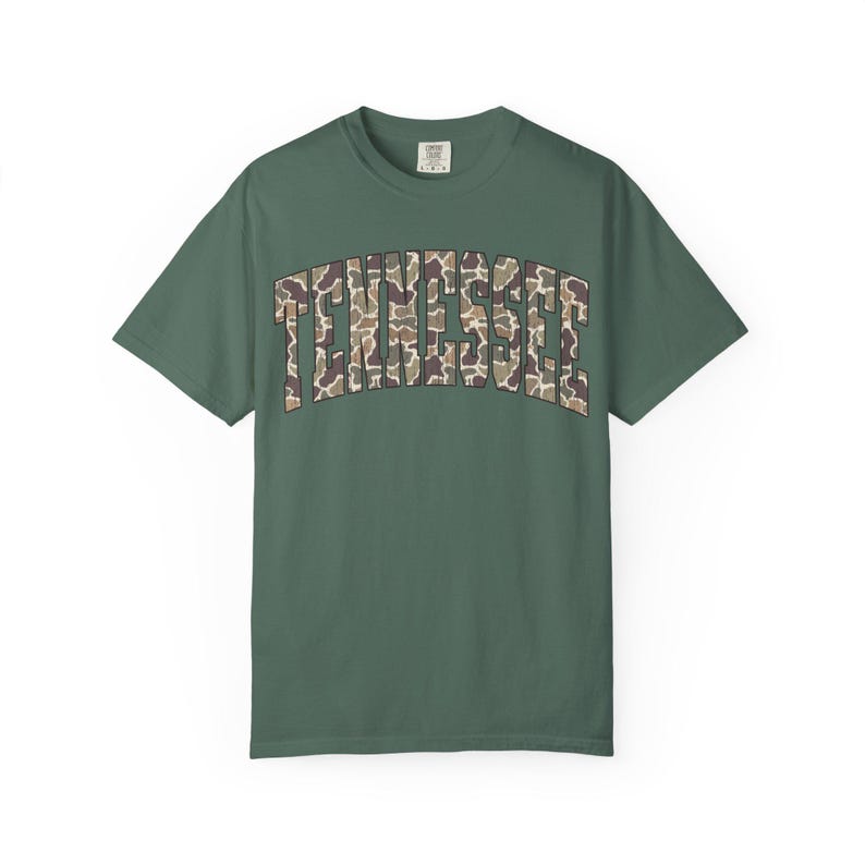 Puede incluir: Camiseta verde con la palabra "TENNESSEE" en un patr&oacute;n de camuflaje en el pecho. La camiseta tiene mangas cortas y cuello redondo. La tela parece ser un material suave y c&oacute;modo.