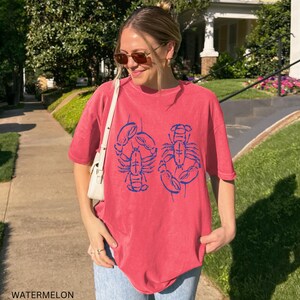 以下が含まれることがあります： 2匹の青いロブスターのイラストがプリントされた、スイカ色のTシャツ。 ゆったりとしたフィット感で、半袖です。 画像の下部には「WATERMELON」の文字がプリントされています。