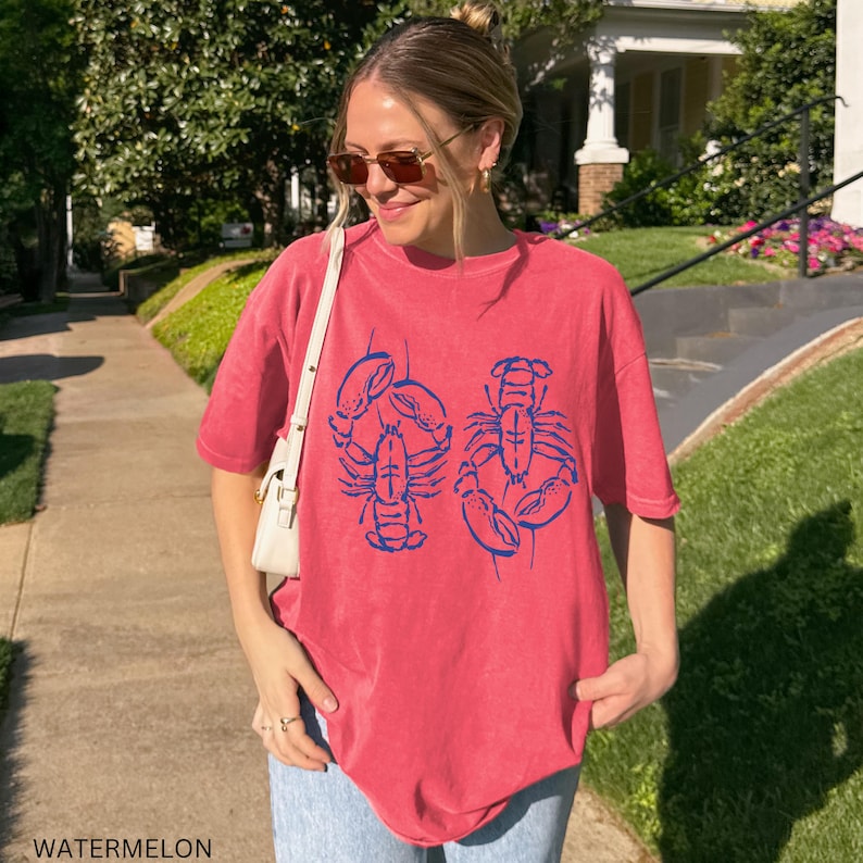 以下が含まれることがあります： 2匹の青いロブスターのイラストがプリントされた、スイカ色のTシャツ。 ゆったりとしたフィット感で、半袖です。 画像の下部には「WATERMELON」の文字がプリントされています。