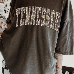 Puede incluir: Camiseta gris oscuro con la palabra "TENNESSEE" en letras grandes en el pecho. Las letras tienen un estampado de camuflaje en tonos verdes, marrones y beige. La camiseta tiene mangas cortas y un corte holgado.