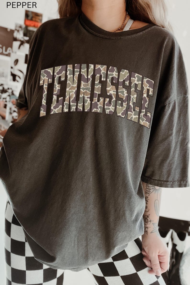 Puede incluir: Camiseta gris oscuro con la palabra "TENNESSEE" en letras grandes en el pecho. Las letras tienen un estampado de camuflaje en tonos verdes, marrones y beige. La camiseta tiene mangas cortas y un corte holgado.