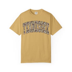 Puede incluir: Camiseta de color amarillo mostaza con la palabra "TENNESSEE" en un estampado de camuflaje en el pecho. La camiseta de manga corta tiene cuello redondo y una etiqueta en la nuca. La camiseta est&aacute; hecha de un material suave y c&oacute;modo.