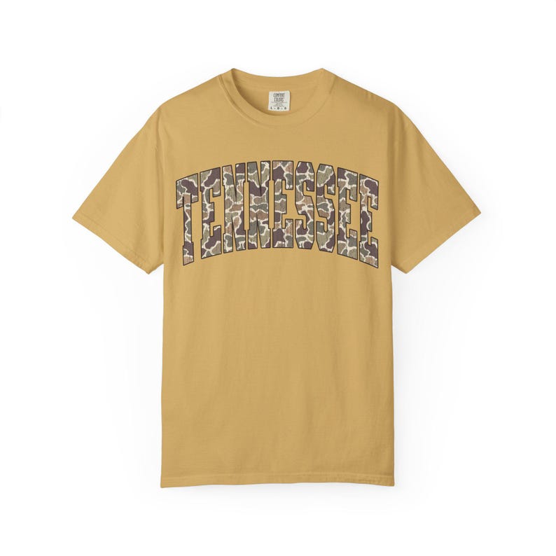 Puede incluir: Camiseta de color amarillo mostaza con la palabra "TENNESSEE" en un estampado de camuflaje en el pecho. La camiseta de manga corta tiene cuello redondo y una etiqueta en la nuca. La camiseta est&aacute; hecha de un material suave y c&oacute;modo.