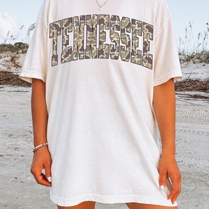 Puede incluir: Camiseta de color marfil con la palabra "TENNESSEE" en un estampado de camuflaje en el pecho. La camiseta tiene mangas cortas y un corte holgado. La palabra "IVORY" est&aacute; impresa en la parte inferior de la imagen.