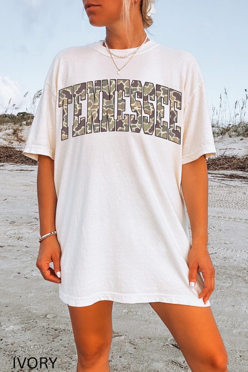 Puede incluir: Camiseta de color marfil con la palabra "TENNESSEE" en un estampado de camuflaje en el pecho. La camiseta tiene mangas cortas y un corte holgado. La palabra "IVORY" est&aacute; impresa en la parte inferior de la imagen.