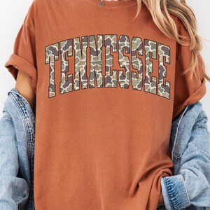 Puede incluir: Una camiseta de color &oacute;xido con la palabra "TENNESSEE" en letras grandes y en bloque en la parte delantera. Las letras est&aacute;n rellenas con un patr&oacute;n de camuflaje. La camiseta tiene mangas cortas y cuello redondo.