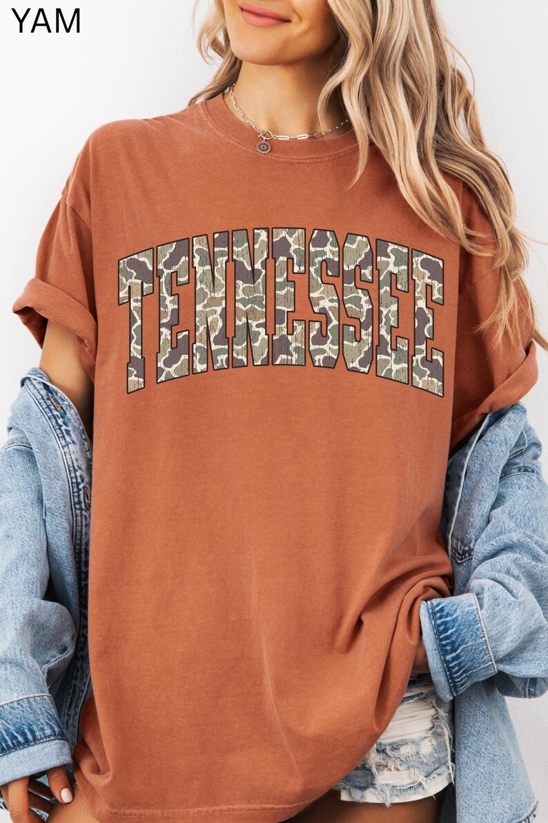 Puede incluir: Una camiseta de color &oacute;xido con la palabra "TENNESSEE" en letras grandes y en bloque en la parte delantera. Las letras est&aacute;n rellenas con un patr&oacute;n de camuflaje. La camiseta tiene mangas cortas y cuello redondo.