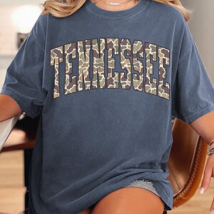 Puede incluir: Una camiseta azul denim con la palabra "TENNESSEE" en letras grandes con un dise&ntilde;o de camuflaje en el pecho. La camiseta tiene mangas cortas y un corte holgado. La palabra "DENIM" est&aacute; en la esquina superior izquierda.