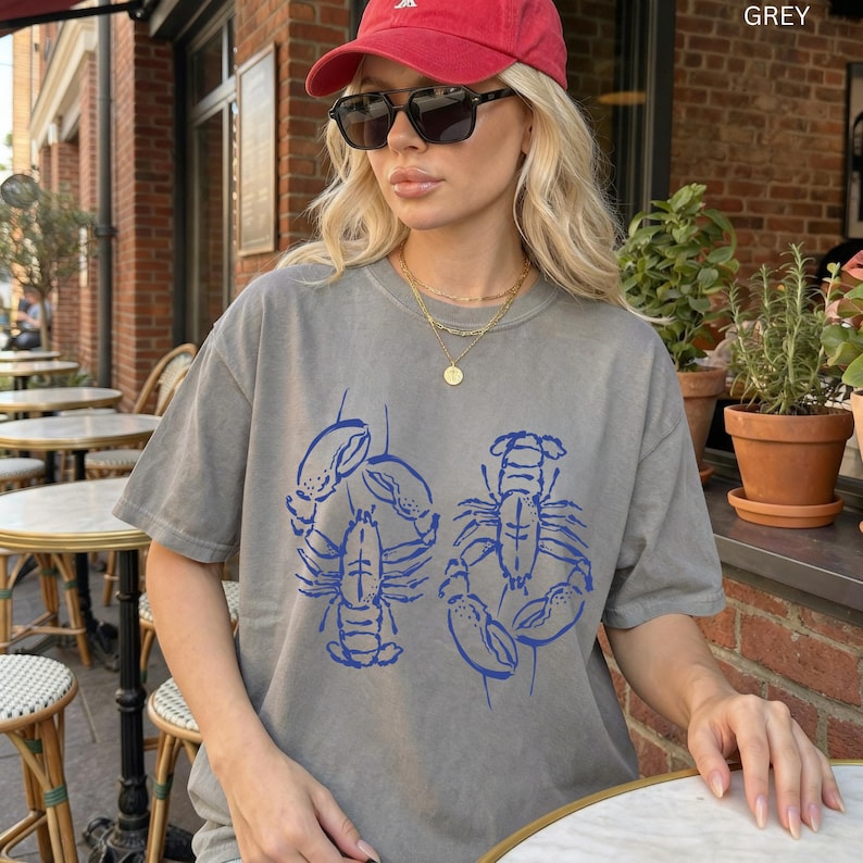 以下が含まれることがあります： 青いロブスターのグラフィックが特徴のグレーのTシャツ。 ゆったりとしたフィット感と半袖です。 右上隅には「GREY」の文字がプリントされています。 赤い野球帽とサングラスを着用しています。