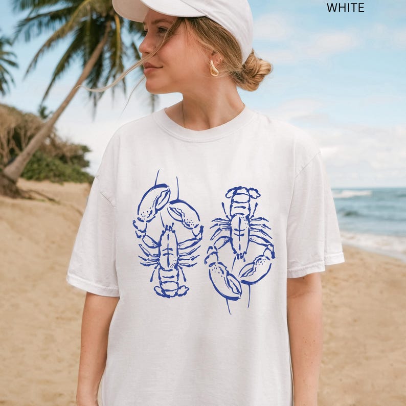 以下が含まれることがあります： 青いロブスターのグラフィックが入った白いTシャツ。デザインは、Tシャツの両側に1つずつ、2つの様式化されたロブスターが特徴です。Tシャツを着ている人は、ヤシの木と海を背景にビーチにいます。