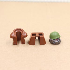 May include: Three plastic toy pieces: a brown saddle, a brown block with two holes, and a green helmet with a gray face mask. The items are likely part of a toy set or building block collection.