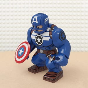 May include: A blue Captain America action figure with a shield. The figure has a blue helmet with a white "A" and white stripes. The shield is red, white, and blue with a white star. The figure is in a crouching position.