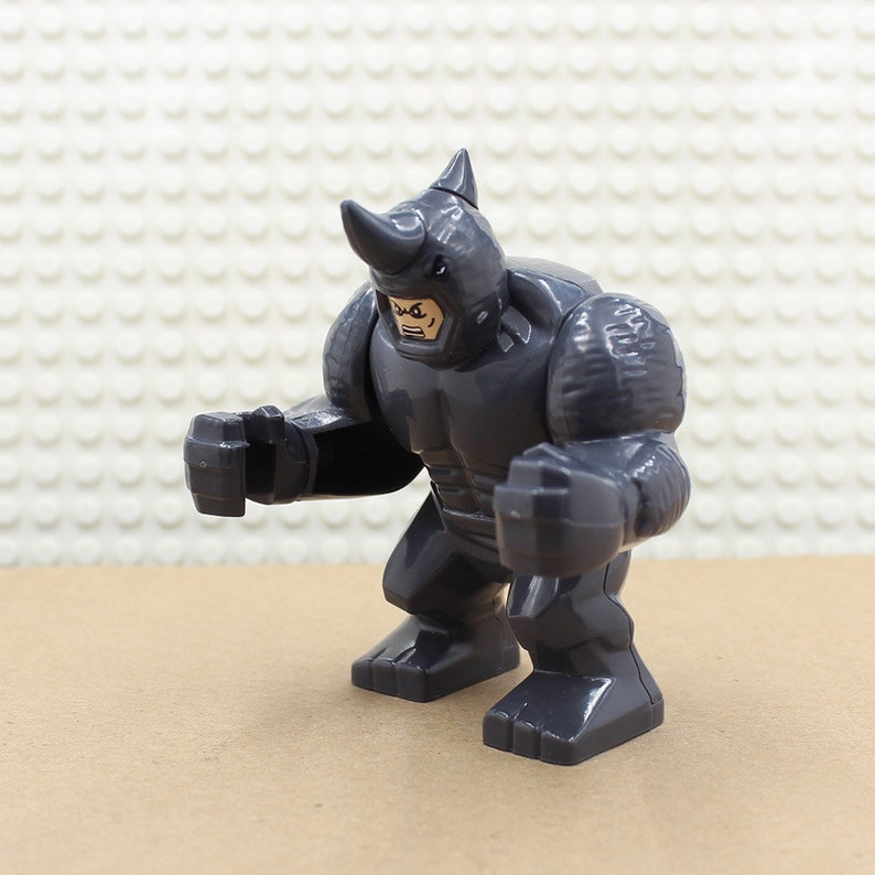 Puede incluir: Una minifigura Lego gris del Rhino de Marvel Comics. La figura tiene una cabeza de rinoceronte gris y un cuerpo gris con armadura texturizada. La figura est&aacute; en posici&oacute;n de combate con los brazos levantados.